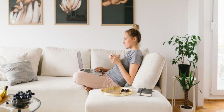 woman sitting on her couch drinking coffee and checking her laptop